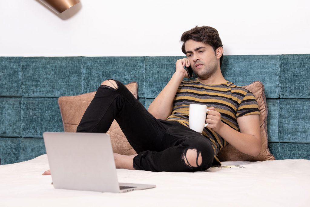 Jaunuolis su kavos puodeliu ir kompiuteriu kalba telefonu namų biure