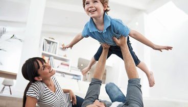 Šviesiame kambarye šeima (tėtis, mama ir vaikas) žaidžia ir spinduliuoja gera, žaisminga emocija